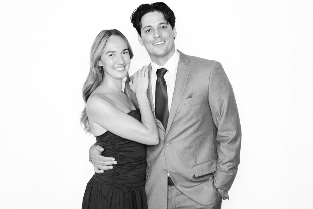 Black-and-white portrait of a smiling couple dressed in formal attire, photographed in a luxury wedding photo booth with a clean studio backdrop.