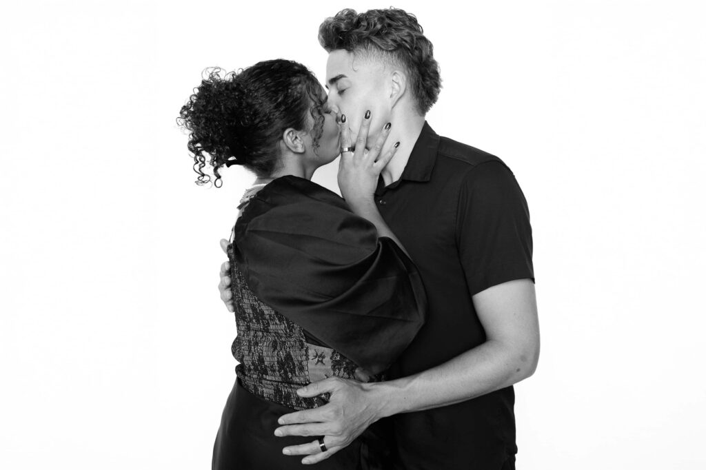 Black-and-white studio-lit portrait of two wedding guests sharing a kiss, photographed in a minimalist white backdrop portrait experience at a luxury reception