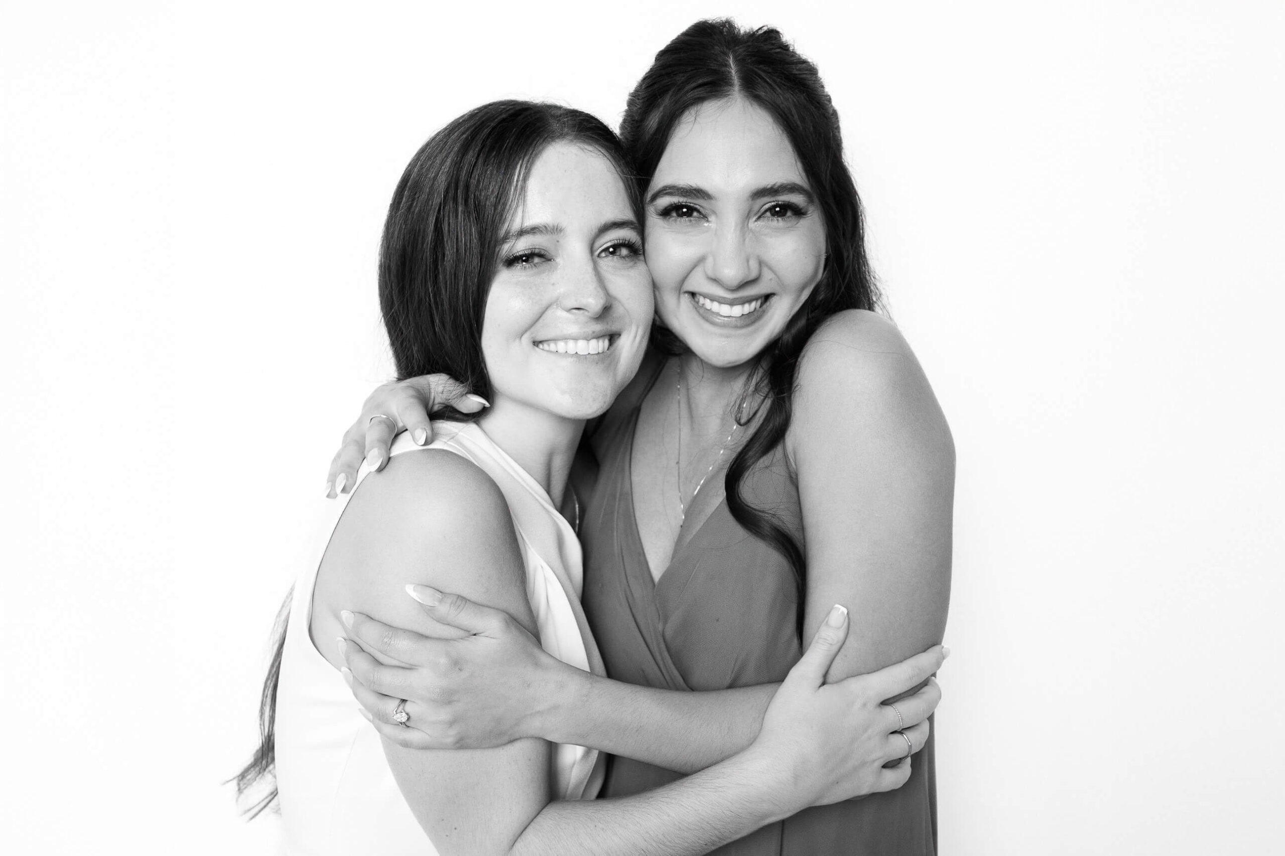 Black-and-white portrait of two women embracing and smiling, photographed in a modern black and white photo booth with soft studio lighting.